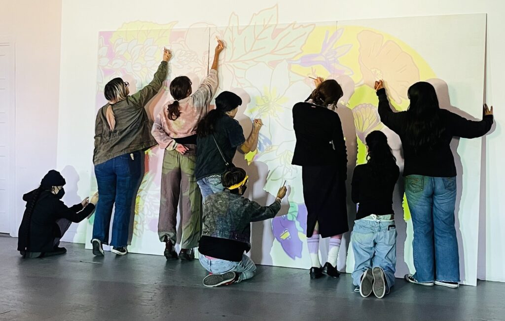 Eight teenagers in jeans and hoodies stand facing a wall with a projection of vibrant, local wildflowers, working together and reaching and tracing the lines to create a large scale mural.
Eight teenagers in jeans and hoodies stand facing a wall with a projection of vibrant, local wildflowers, working together and reaching and tracing the lines to create a large scale mural.