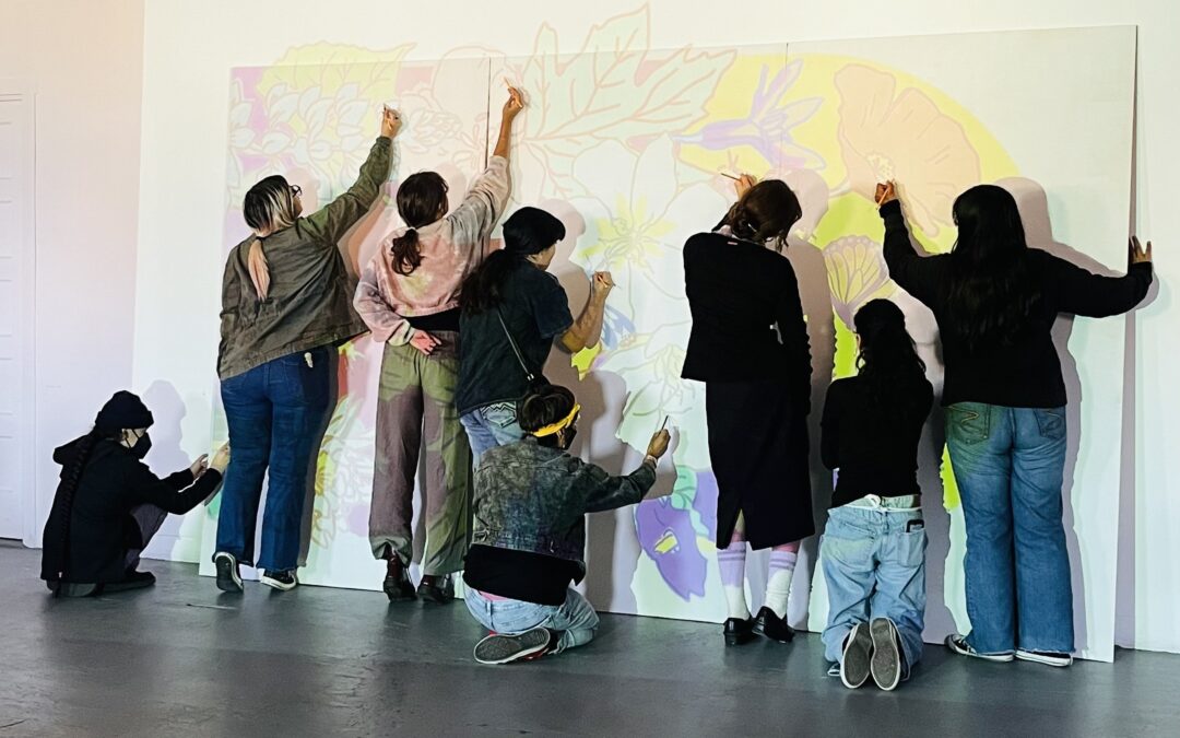 Eight teenagers in jeans and hoodies stand facing a wall with a projection of vibrant, local wildflowers, working together and reaching and tracing the lines to create a large scale mural. Eight teenagers in jeans and hoodies stand facing a wall with a projection of vibrant, local wildflowers, working together and reaching and tracing the lines to create a large scale mural.
